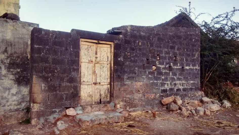An old, weathered stone building with a faded wooden door, set against a dry, rural landscape in an Indian village. Crumbling rocks and dry brush surround the structure, suggesting abandonment or decay.