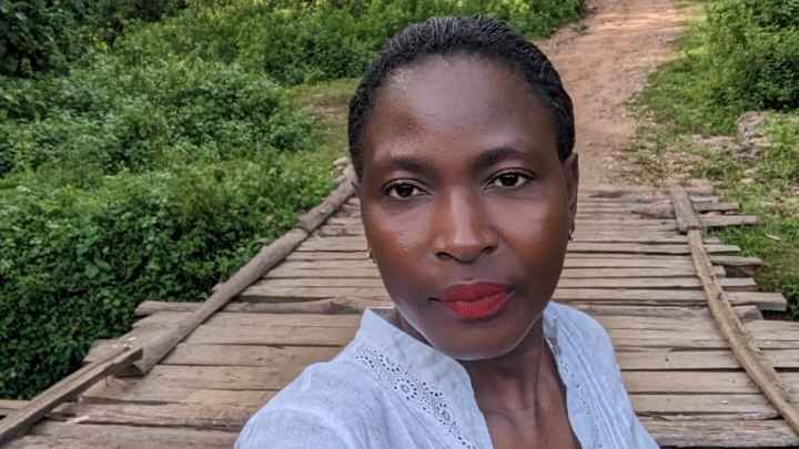 Leonida Odongo is a Black woman with Black hair that is pulled back into an updo. She wears bright red lipstick and a white linen blouse with a small pair of gold earrings. Behind her is a wooden and dirt path and greenery.