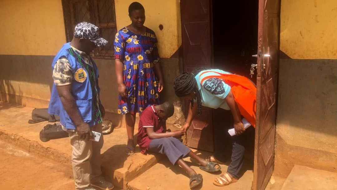 From left to right: A community health worker in a blue vest and khaki pants looks on as Bless, a teenage boy in a red shirt, blue pants, and sandals greets Nain Yuh, who's in a light blue shirt. Behind Bless is his mother in a colorful blue dress.