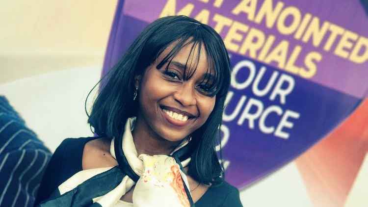 Bolutife Asake smiles in front of a large poster. She has brown skin and black shoulder-length hair with bangs. She wears a black long-sleeved top and black and cream-colored scarf.