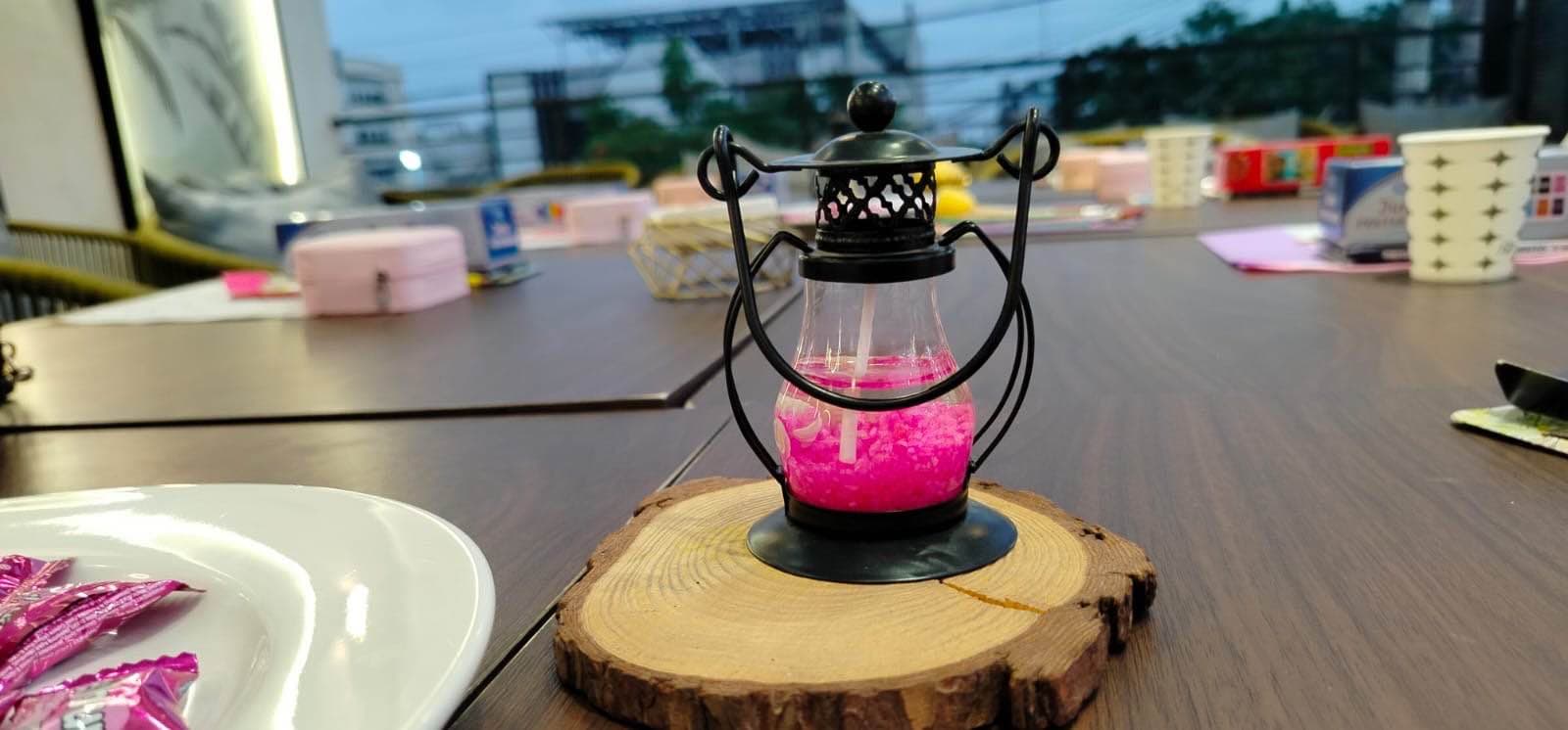 A small pink lantern with a black metal frame sits on a round wooden surface outdoors. The background is softly blurred, with greenery and an open outdoor setting visible behind it.