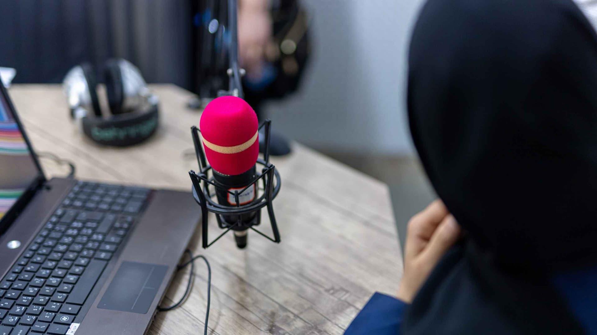 A woman radio announcer in a black hijab speaks over a red microphone