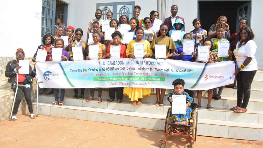 Women with varied disabilities, dressed in a beautiful rainbow of colours and smiling, stand for a family picture after a workshop on GBV.